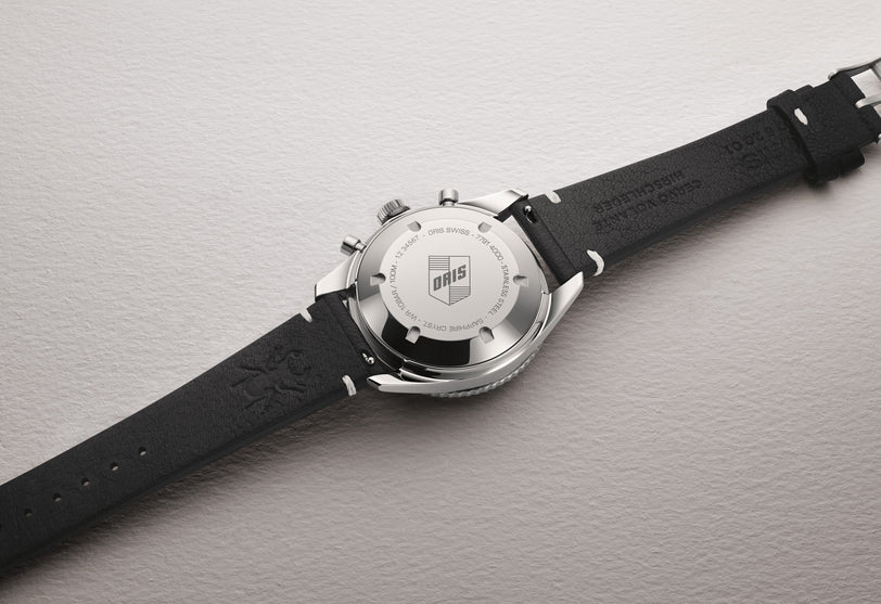 A close-up of the Oris Divers Sixty-Five Chronograph’s stainless steel back and black leather strap, displayed on a light textured surface.