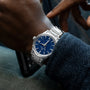 A person wearing a Grand Seiko Automatic GMT SBGM239 Limited Edition For HODINKEE with a twilight blue dial and date display is seen from the side while holding a smartphone.
