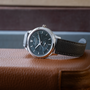A Chopard L.U.C XPS 1860 Officer Limited Edition For Hodinkee with a white gold case and guilloché dial sits on a brown leather surface. This silver Chopard watch has a black textured band and displays the day, date, and time.