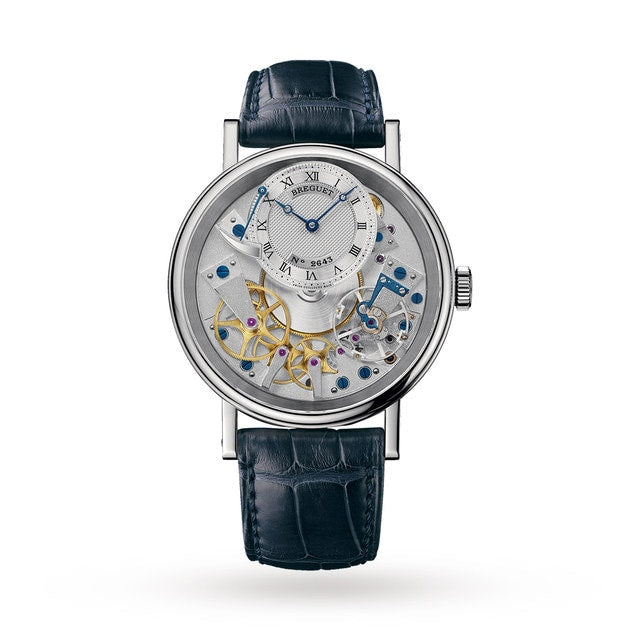 The Breguet Tradition 7057 is a luxury wristwatch with a black leather strap, white gold case, and an exposed hand-wound movement that showcases intricate gears beneath a compact time display.