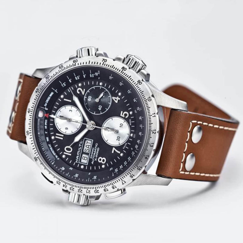 The Hamilton Khaki Aviation X-Wind Auto Chrono is a silver chronograph wristwatch featuring a black dial, three subdials, and a brown leather strap with metal rivets, inspired by classic pilot’s timepieces, shown on a white background.