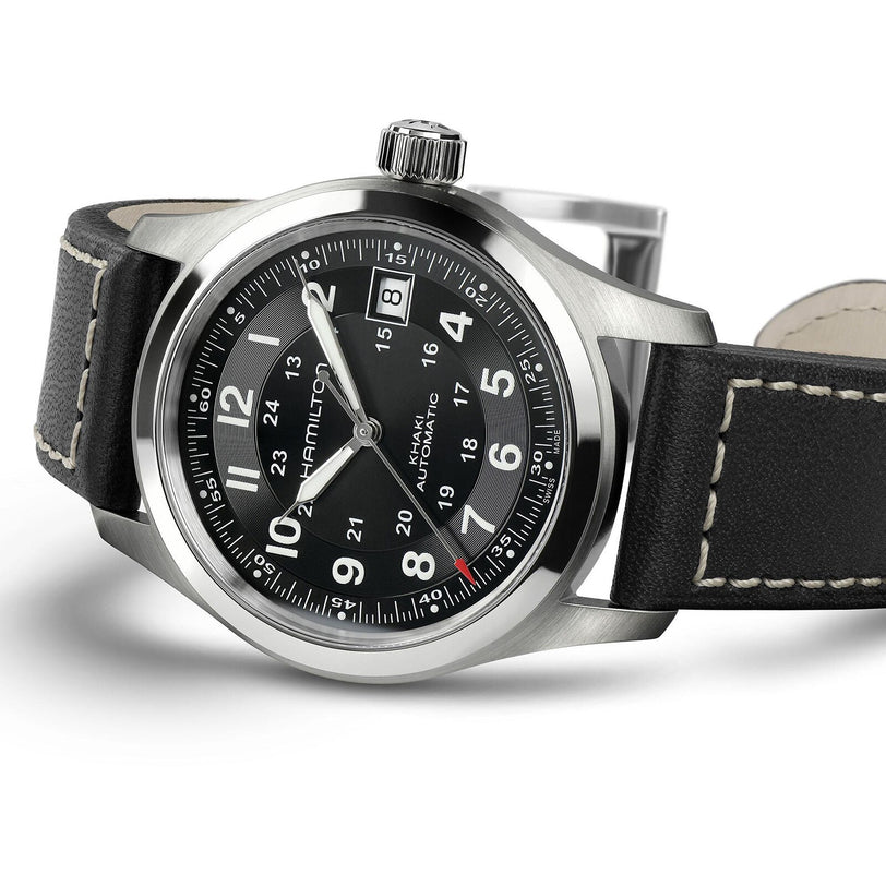 The Hamilton Khaki Field Auto is a rugged, military-inspired watch with a black dial, white numerals, and a black leather strap with white stitching. The time shown is 10:10.