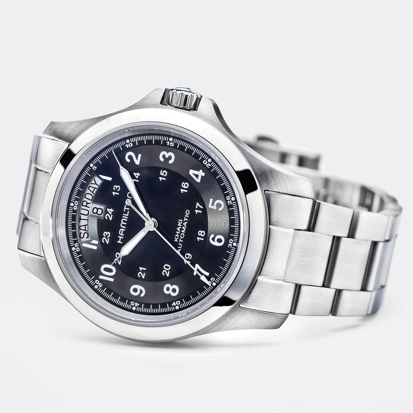 The Hamilton Khaki Field King Auto is a stainless steel wristwatch with a black dial, white numerals, military time markings, metal link bracelet, and left-side day/date display.