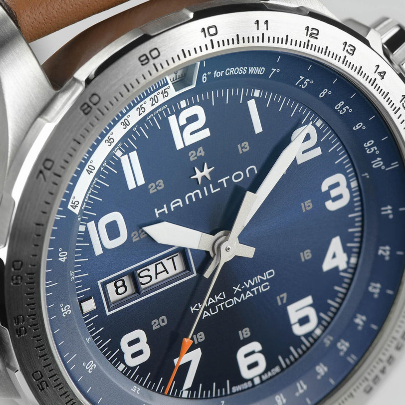 Close-up of a Hamilton Khaki Aviation X-Wind Day Date Auto watch with a blue dial, SuperLumiNova® numerals, date display showing 8 SAT, stainless steel case, and part of a brown strap visible.