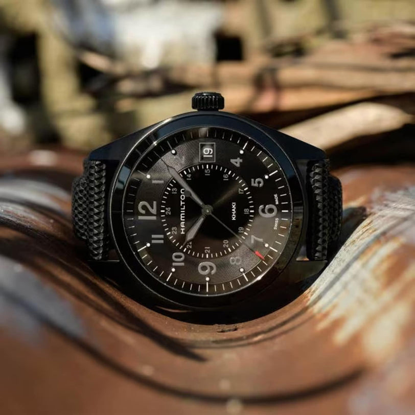 The Hamilton Khaki Field Quartz wristwatch, featuring a textured black strap and unique design, is showcased outdoors on a rusted, wavy metal surface.
