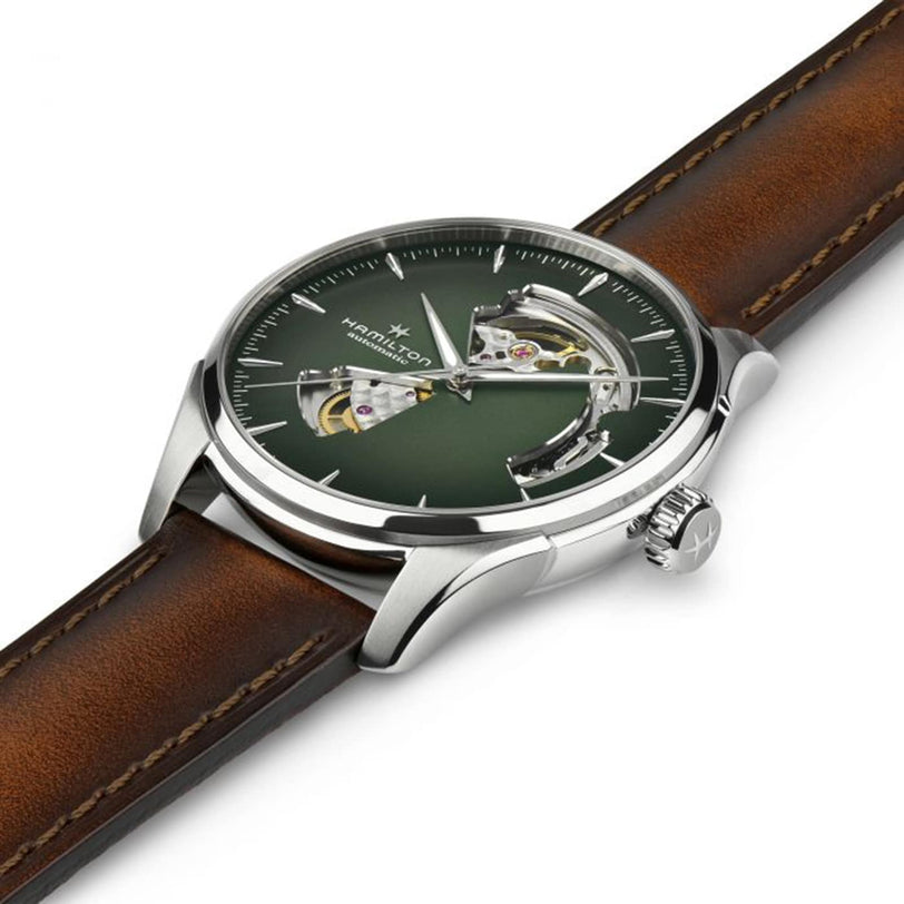 A Hamilton Jazzmaster Open Heart Auto wristwatch featuring a green smoked dial, silver case, exposed gears, and a brown leather strap is shown angled on a white background.