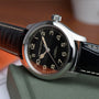 A Hamilton Khaki Field Murph 38mm watch with a black dial, white numerals, automatic movement, and black leather strap sits on a green textured surface.