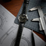 A Hamilton Khaki Field Murph 38mm with an 80-hour power reserve and black leather strap rests on a notebook with watch sketches, next to a caliper, ruler, and pen on a wooden desk.
