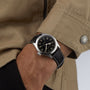 Someone in a tan jacket and black pants is wearing the Hamilton Khaki Field Murph 38mm, which has a silver case, black leather strap, black dial, and offers an 80-hour power reserve.