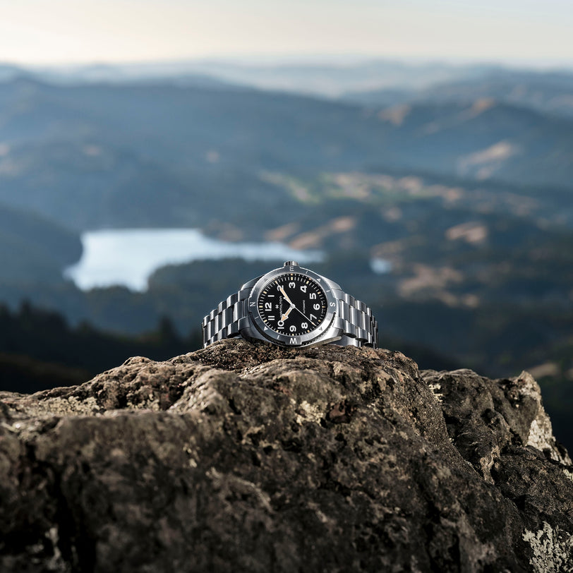 The Hamilton Khaki Field Expedition Auto, featuring a black dial and stainless steel case, rests atop a rocky surface with a blurred mountain landscape and lake in the background.