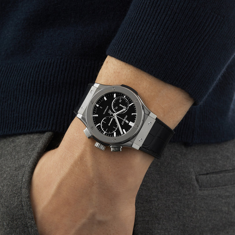 A person in a navy sweater and gray pants wears the Hublot Classic Fusion Chronograph Titanium, featuring a black dial, black leather strap, titanium case, and self-winding movement.