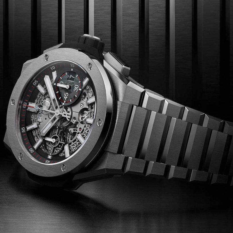 The Hublot Big Bang Integrated Titanium wristwatch features a visible mechanical movement, black and red accents, and a metal link bracelet, resting on a brushed metal surface that reflects its bold titanium design.