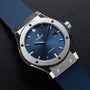 The Hublot Classic Fusion Titanium Blue features a blue dial and strap, silver bezel, and automatic movement, displayed on a black surface.