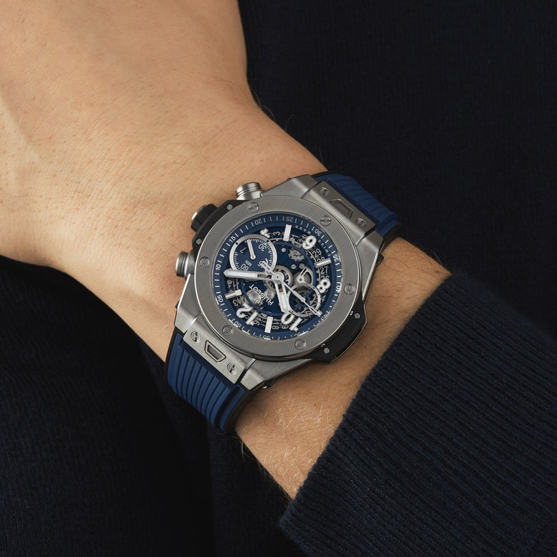 Someone wearing a Hublot Big Bang Unico Titanium Blue—a large chronograph wristwatch with a skeleton dial, visible gears, blue rubber strap, and bold titanium case—on their left wrist.