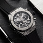 A close-up of the Hublot Big Bang Unico Titanium, highlighting its skeleton chronograph flyback movement, black rubber strap, and titanium case on a dark surface beside a pen and metallic object.