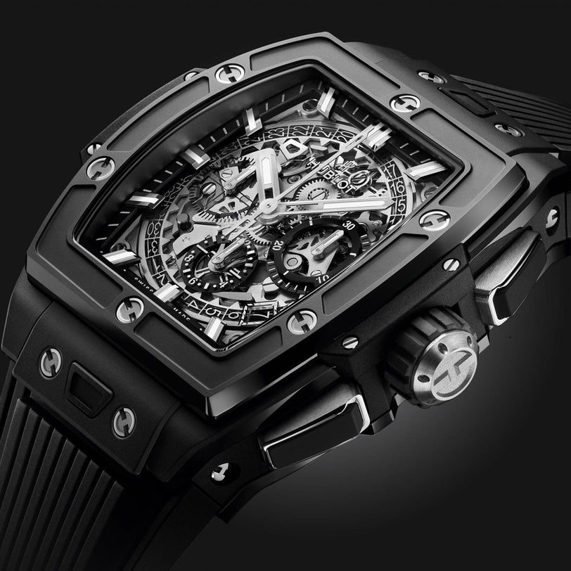 Close-up of the Hublot Spirit Of Big Bang Black Magic, a luxury black wristwatch with a square ceramic case, skeleton dial showing intricate gears, multiple subdials, metal screws, and a textured rubber strap against a dark background.