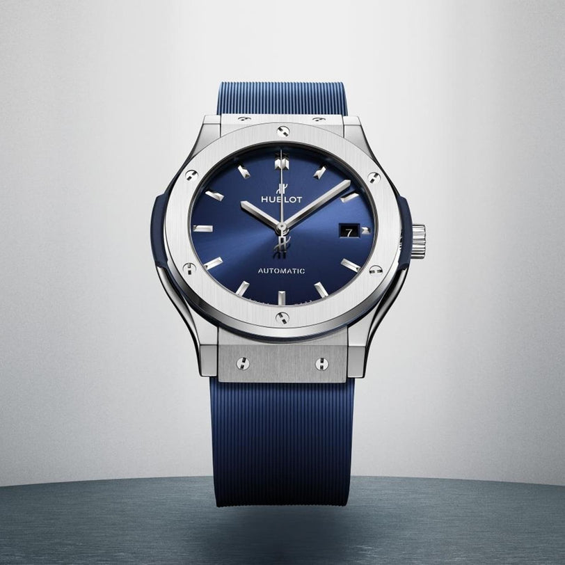 The Hublot Classic Fusion Titanium Blue wristwatch, featuring a blue dial and blue strap, is displayed upright on a grey surface.