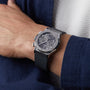A person wearing a Hublot Classic Fusion Racing Grey Chronograph Titanium watch with a black strap and a dark blue jacket over a white shirt.