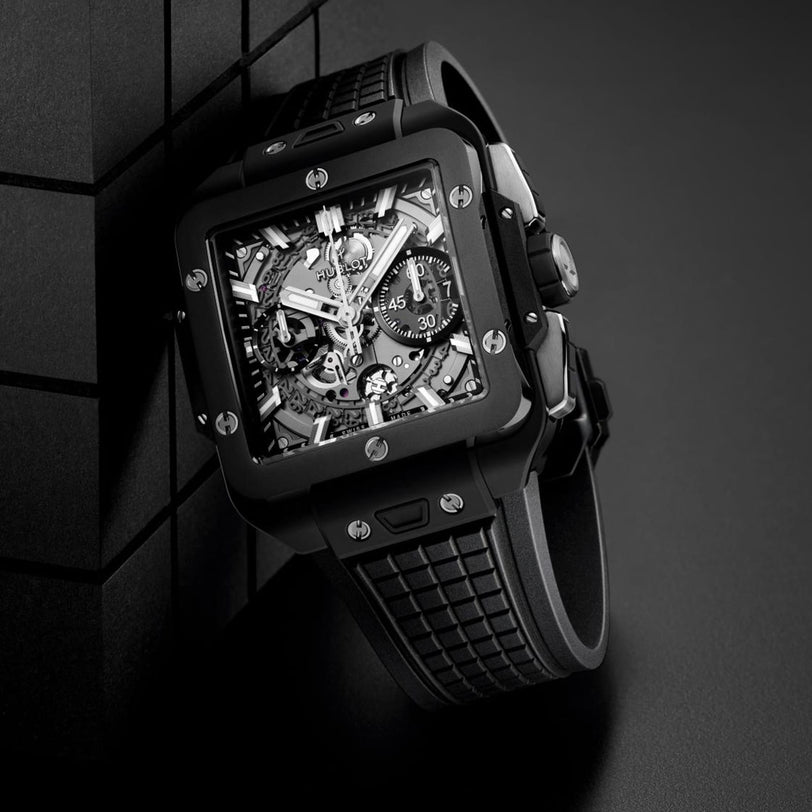 The Hublot Square Bang Unico Black Magic wristwatch, featuring a sapphire crystal skeleton dial and rubber strap, rests against a black geometric surface on a dark background.