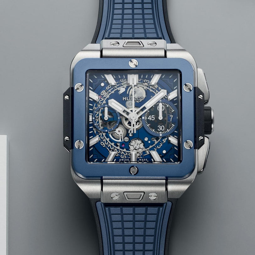 The Hublot Square Bang Unico Titanium Blue Ceramic wristwatch features a HUB1280 UNICO movement, blue rubber strap, sapphire crystal dial, and chronograph dials, shown against a gray background.