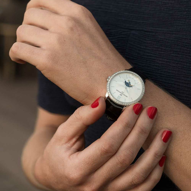 Person with red nail polish adjusts a round Girard-Perregaux 1966 Moon Phases wristwatch with a white face, using their other hand.