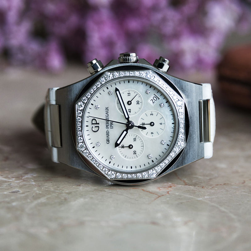 The Girard-Perregaux Laureato Chronograph 38, with an octagonal bezel, diamond-set accents, and date display, is photographed on marble with purple flowers in the background.