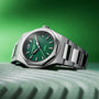 The Girard-Perregaux Laureato 42, featuring a silver case, Sunray Green dial, and metal band, is displayed on a gray wavy surface with a green background.