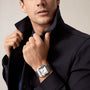 A man in a dark jacket adjusts his collar, showcasing a Cartier Santos de Cartier silver wristwatch with a square face on his wrist.