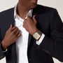 A person in a dark suit and white shirt adjusts their jacket, revealing a Cartier Santos de Cartier Chronograph watch with a metallic band on their wrist.