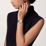 A woman in a sleeveless navy cable-knit top touches her face, wearing a Cartier Santos-Dumont watch with an 18K rose gold square face.