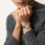 A person in a gray sweater holds their hands near their face, showcasing a Cartier Santos-Dumont watch with an 18K rose gold case and black leather strap.