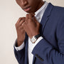 A person in a blue suit adjusts the collar of a white shirt, wearing a Ballon Bleu de Cartier watch by Cartier with a black leather strap on their wrist.