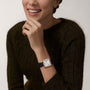 A woman in a dark green sweater smiles and touches her chin, showcasing a Ballon Bleu de Cartier watch on her left wrist—an elegant timepiece by Cartier.