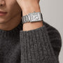 A person in a grey knit sweater showcases the Cartier Tank Américaine watch, featuring a rectangular silvered flinqué dial with Roman numeral markers on a metal band.