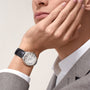 A person in a gray suit rests their chin on their hand, showcasing a Cartier Ballon Bleu de Cartier watch with a white face and black alligator leather strap.