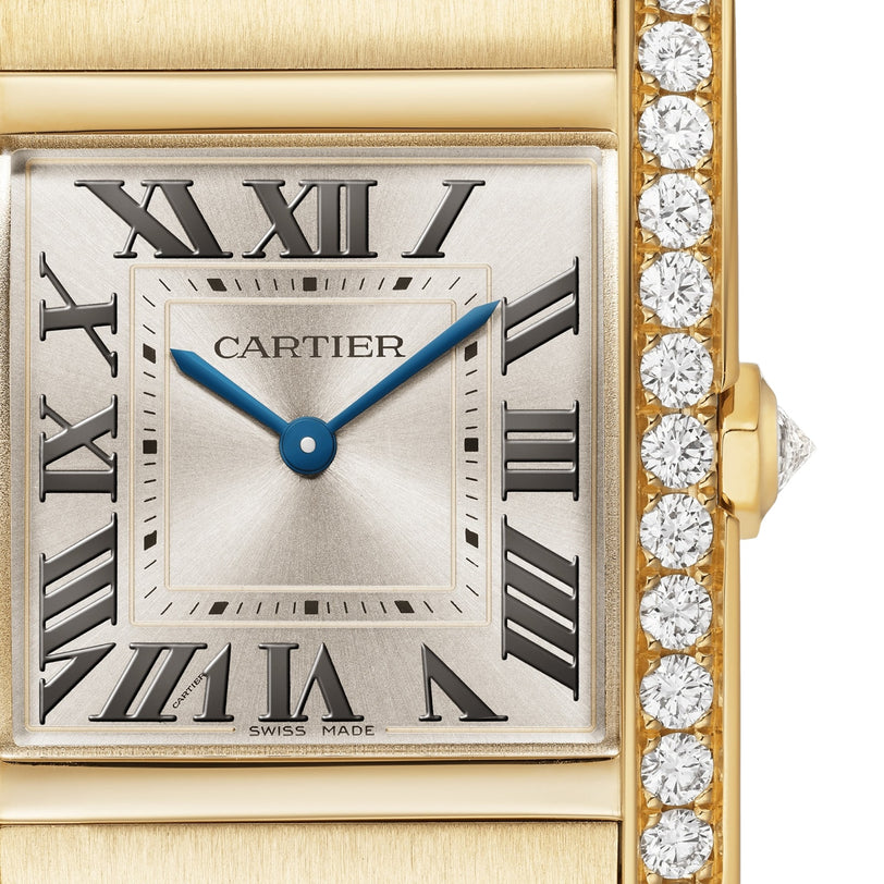 Close-up of a Cartier Tank Américaine watch face with Roman numerals, blue hands, a yellow gold case, and a row of brilliant-cut diamonds along the edge.