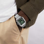 A person displays form precision wearing Cartier’s Santos de Cartier watch with a silver case, green dial, and Roman numerals, styled with a white shirt, olive jacket, and beige pants, one hand tucked into their pocket.