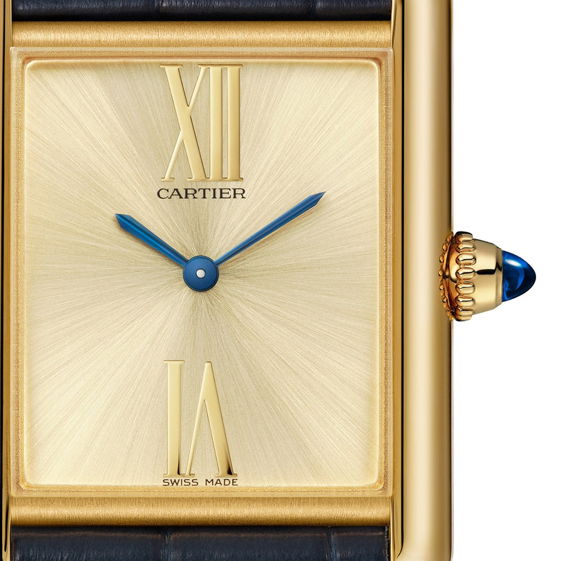 Close-up of a Cartier Tank Américaine watch face featuring a gold rectangular case, blue hands, prominent Art Deco Roman numerals at 12 and 6, and a signature blue cabochon crown.