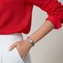 A person in a red long-sleeve blouse and white pants, hand in pocket, showcases the Cartier Panthère de Cartier watch with a sleek steel case and rectangular face.
