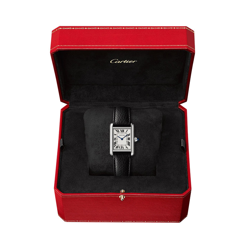 A Cartier Tank Must de Cartier watch with a rectangular face, black leather strap, and beaded crown is displayed on a black cushion inside a red Cartier box.
