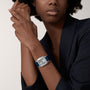 A person in a dark blazer showcases a Cartier Santos-Dumont watch with a silver case and blue strap on their left wrist, holding their right hand near their face.