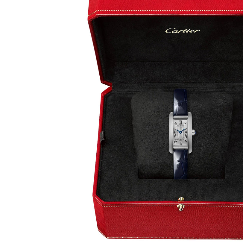 A Cartier Tank Américaine watch with a blue leather strap rests on a black cushion inside an open red Cartier box.