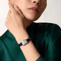 Wearing a green satin top, a person showcases the Cartier Tank Américaine watch—rectangular silver case and dark strap—on their left wrist, with their hand thoughtfully positioned near their chin.