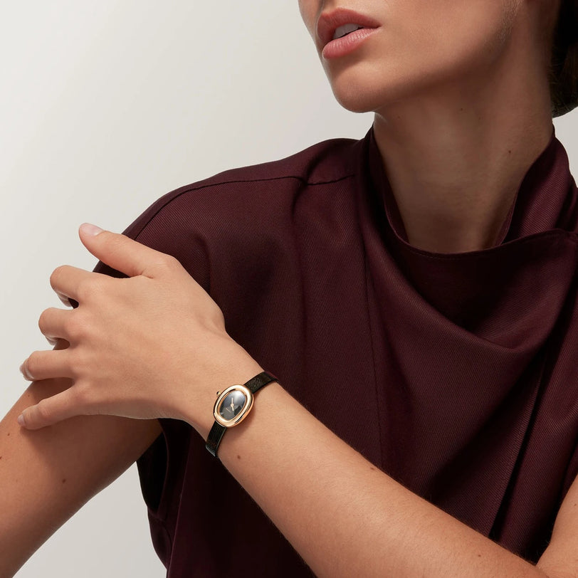 A woman in a burgundy top touches her shoulder while wearing a Cartier Baignoire gold and black wristwatch.