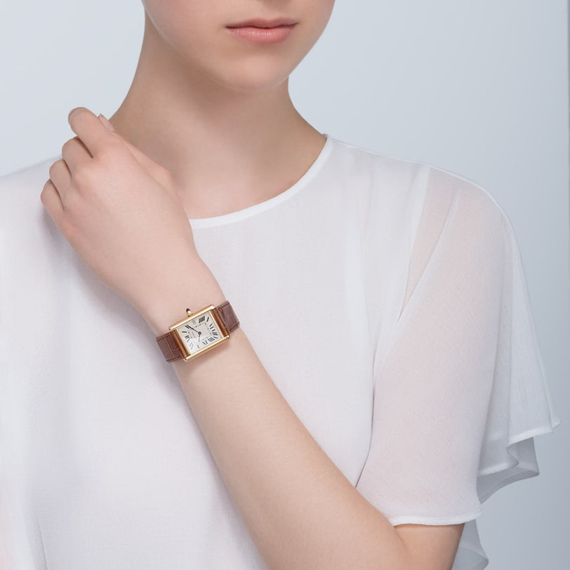 A person wearing a white sheer blouse shows off a Cartier Tank Louis Cartier watch with a square face and brown leather strap on their left wrist.