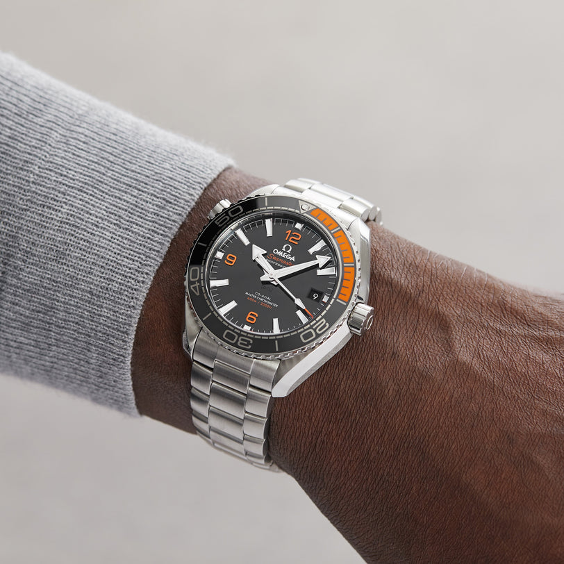 A person wearing an OMEGA Seamaster Planet Ocean 600M with a black and orange dial, black ceramic bezel, stainless steel band, and a gray long-sleeve shirt.