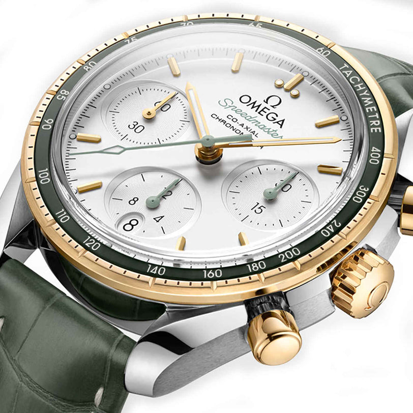 Close-up of the OMEGA Speedmaster 38 wristwatch with a white dial, green bezel, gold details, and green leather strap. Features three subdials, a tachymeter scale, and a Co-Axial movement inspired by lunar missions.