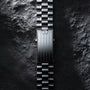 The OMEGA Speedmaster Moonwatch Professional’s metal bracelet, featuring the iconic logo on its clasp, is displayed vertically on a textured, stone-like surface—a tribute to its legendary legacy.