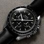 The OMEGA Speedmaster Moonwatch Professional, featuring a black dial with three sub-dials, tachymeter bezel, and black leather strap with 3861 calibre movement, is photographed on a textured gray surface.