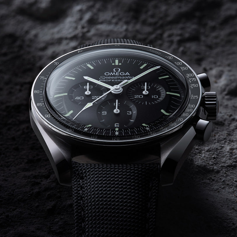 A close-up of the OMEGA Speedmaster Moonwatch Professional with a black dial and fabric strap, powered by the 3861 calibre movement, displayed on a textured dark surface.